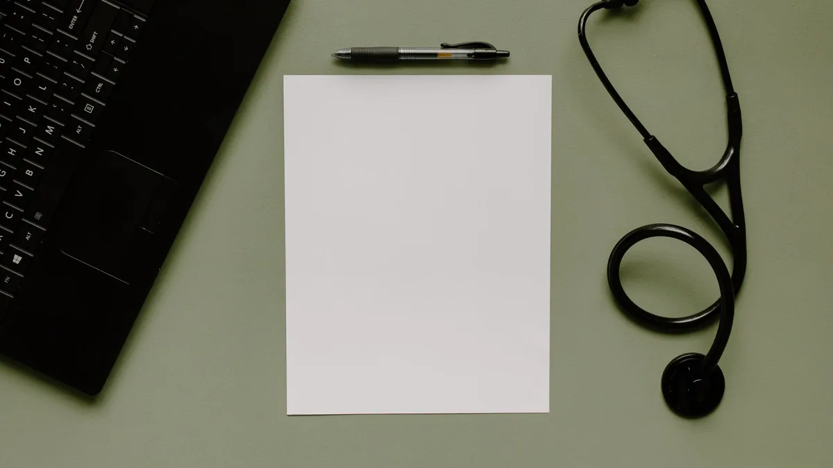 Medical workspace featuring a blank paper, stethoscope, and laptop from above.