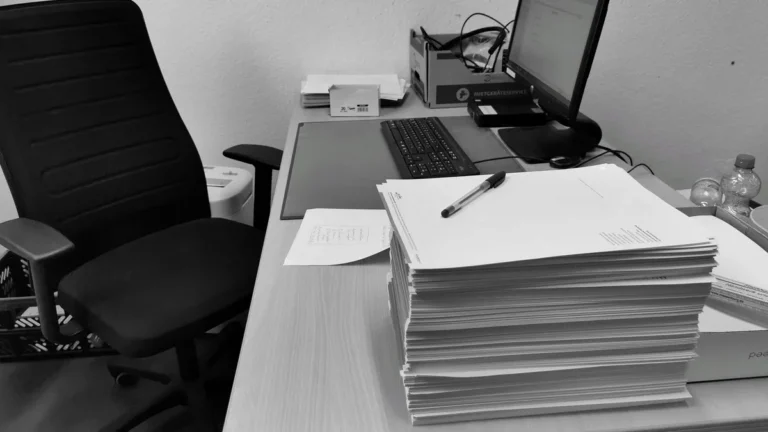 Black and white image of a cluttered office desk with a computer, chair, and paperwork.